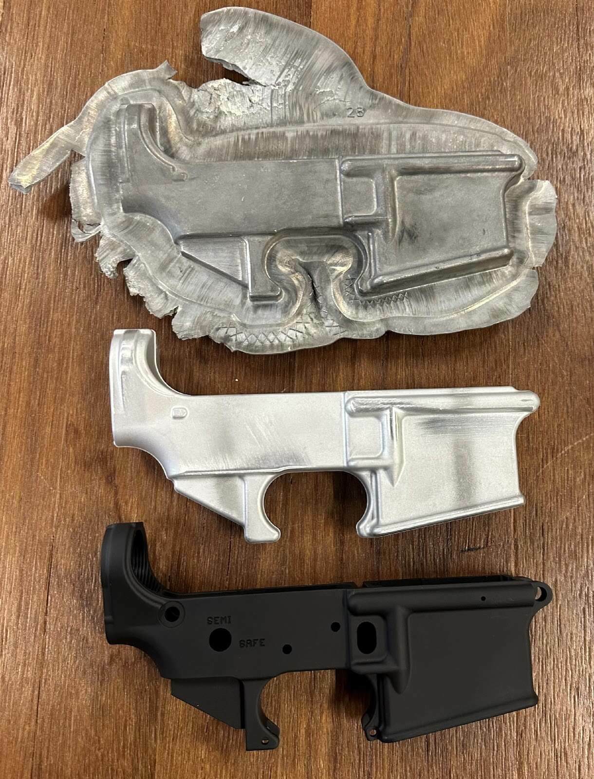 Three firearm receiver components in various materials and finishes on a wooden surface.