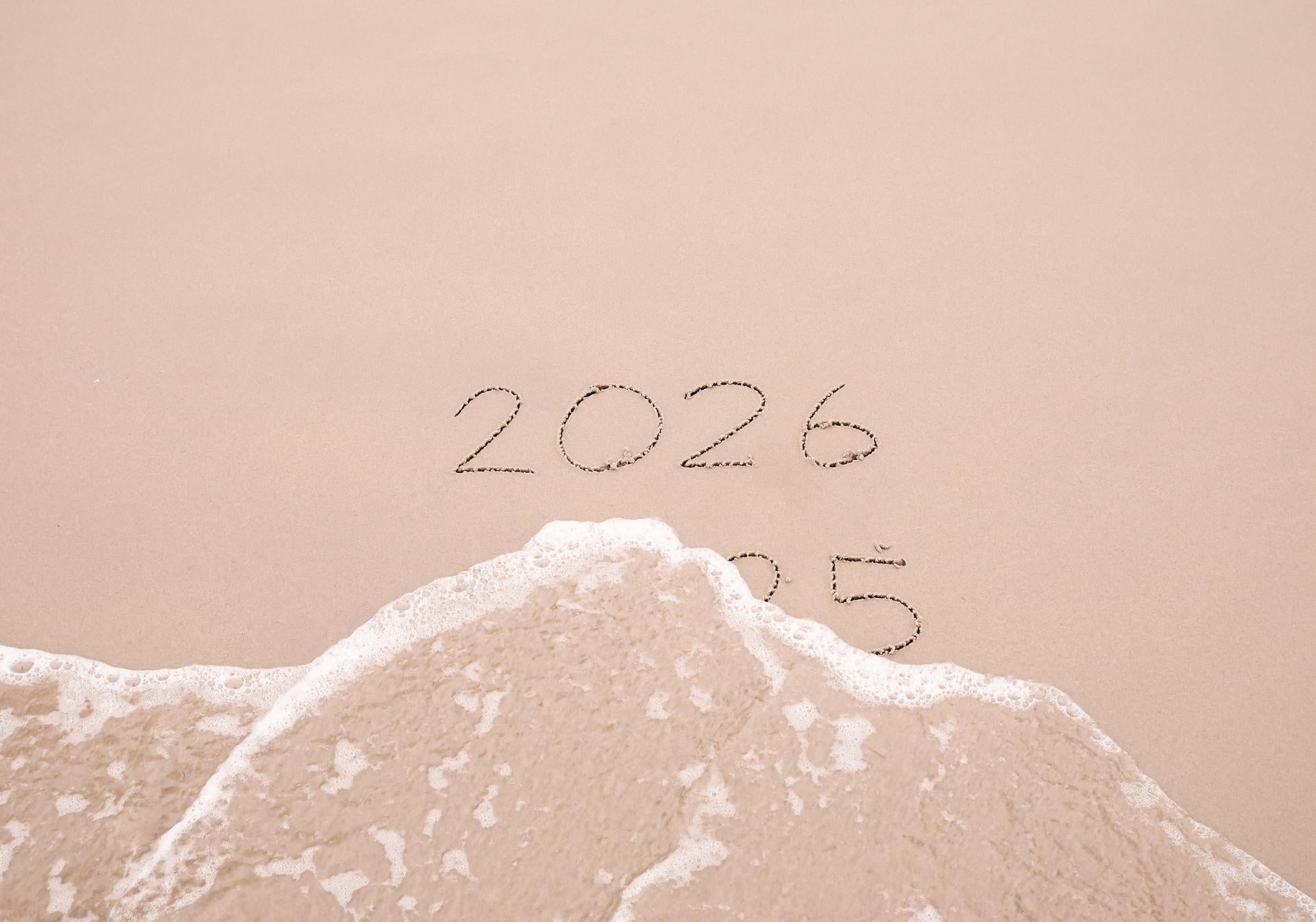 Handwritten inscription 2025 and 2026 with ocean waves on a sand beach. The concept of Goodbye 2025 and Happy New Year 2026