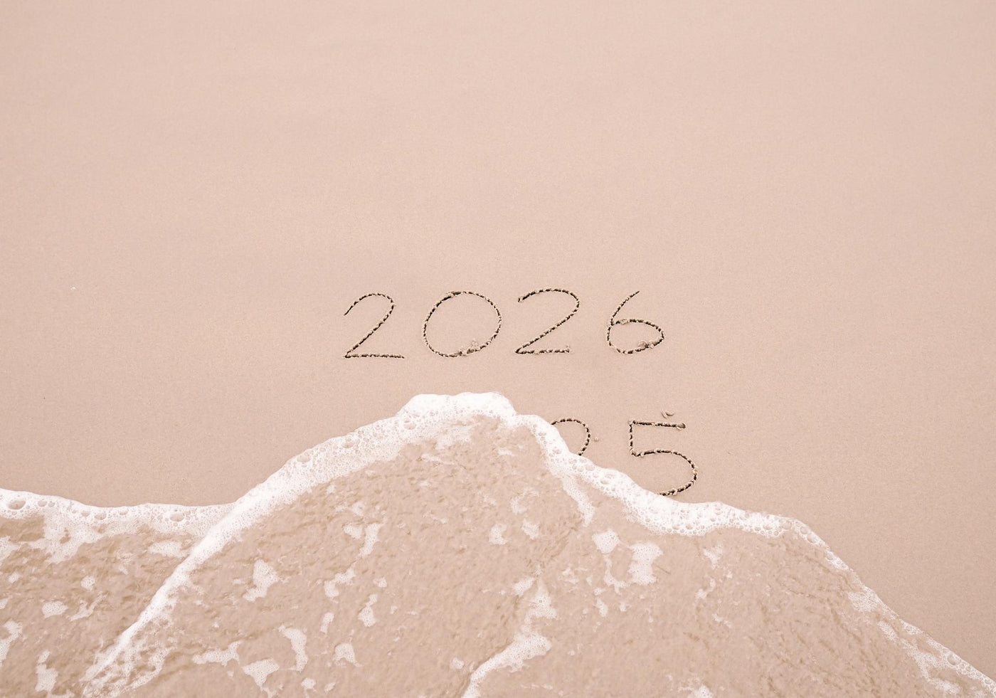 Handwritten inscription 2025 and 2026 with ocean waves on a sand beach. The concept of Goodbye 2025 and Happy New Year 2026