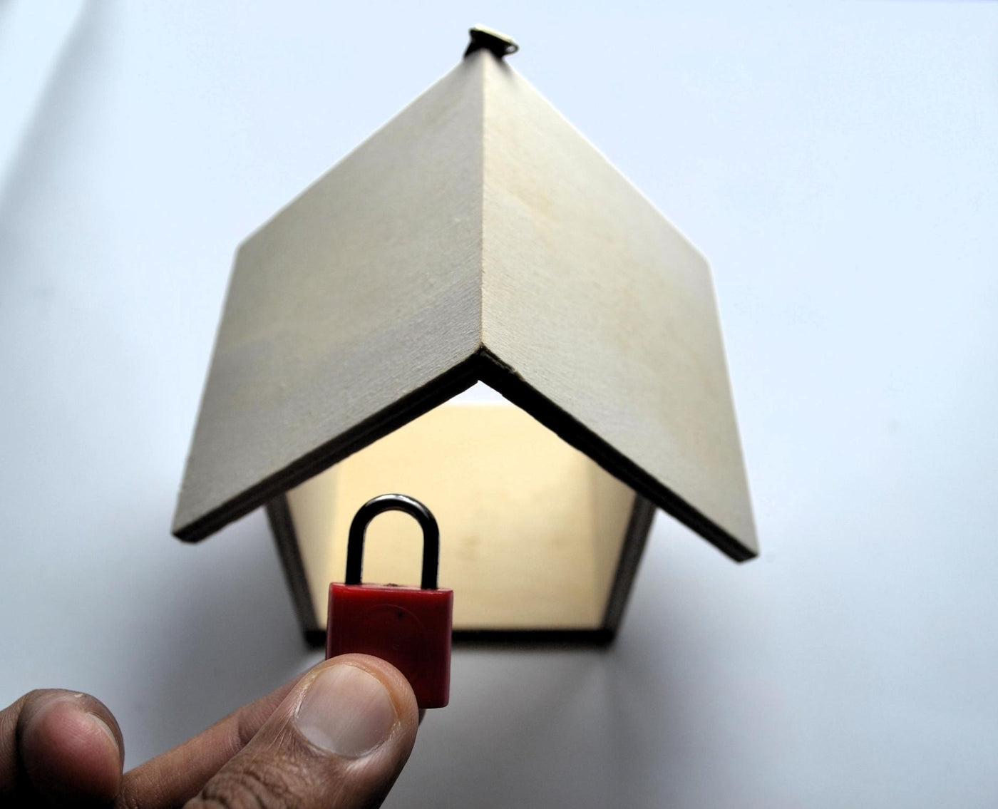 Hand holding red padlock beneath wooden house model on white background symbolizing home safety, property protection, and secure living for lifestyle, insurance, and editorial stock visuals.