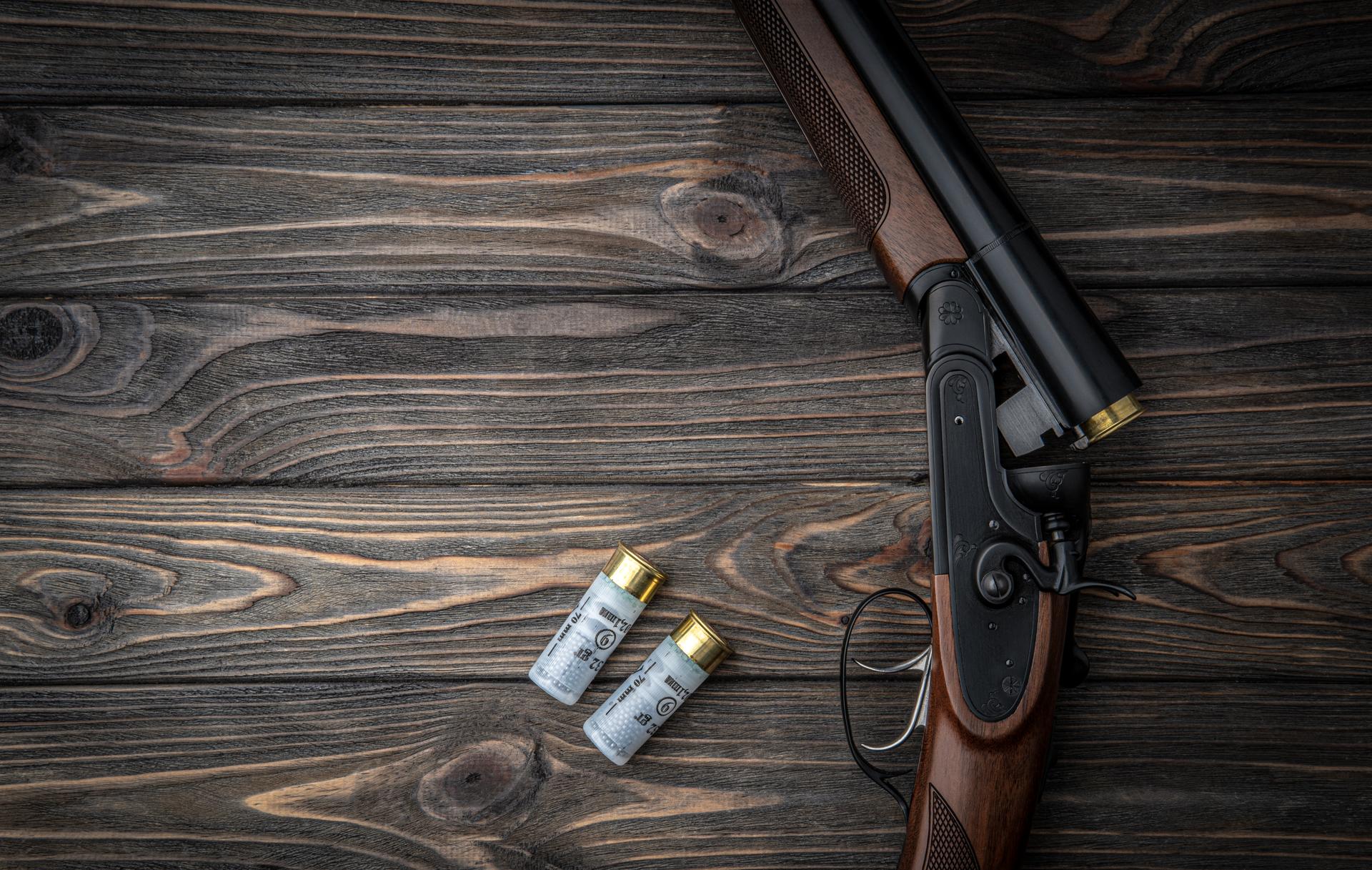 Opened double-barreled hunting rifle on a wooden table. Shotgun ammo. Dark brown wooden back.