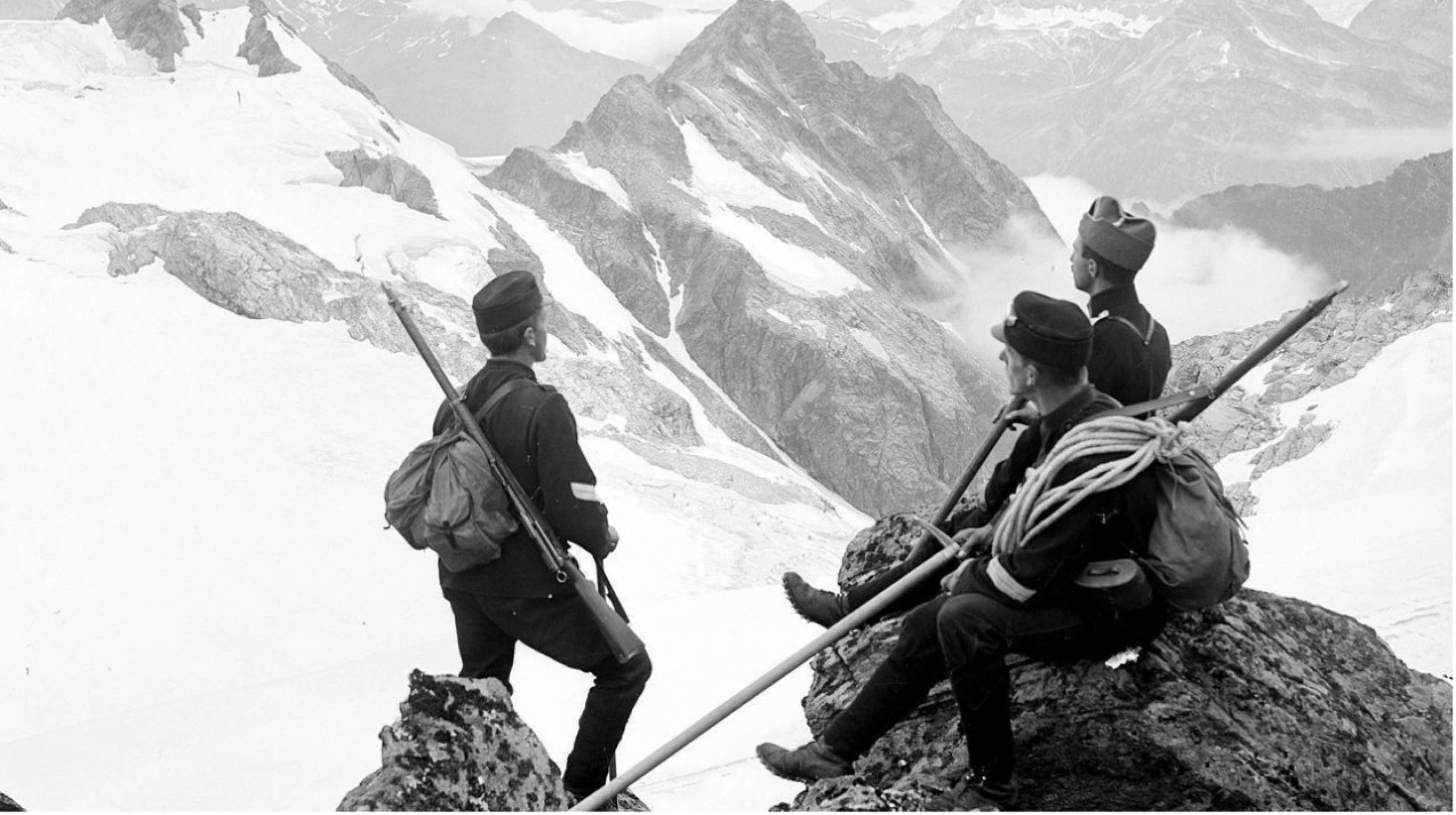 Three climbers resting on a rocky outcrop with mountains in the background.