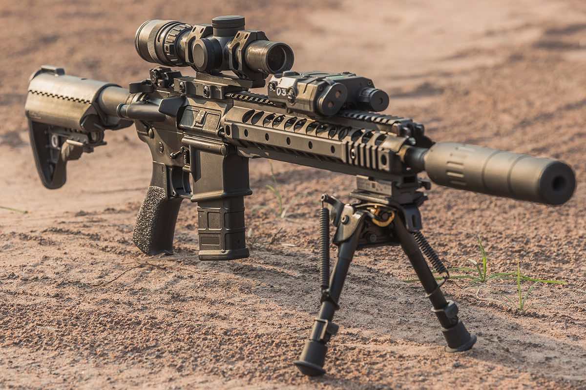 A tactical rifle with a scope and bipod on a sandy surface.